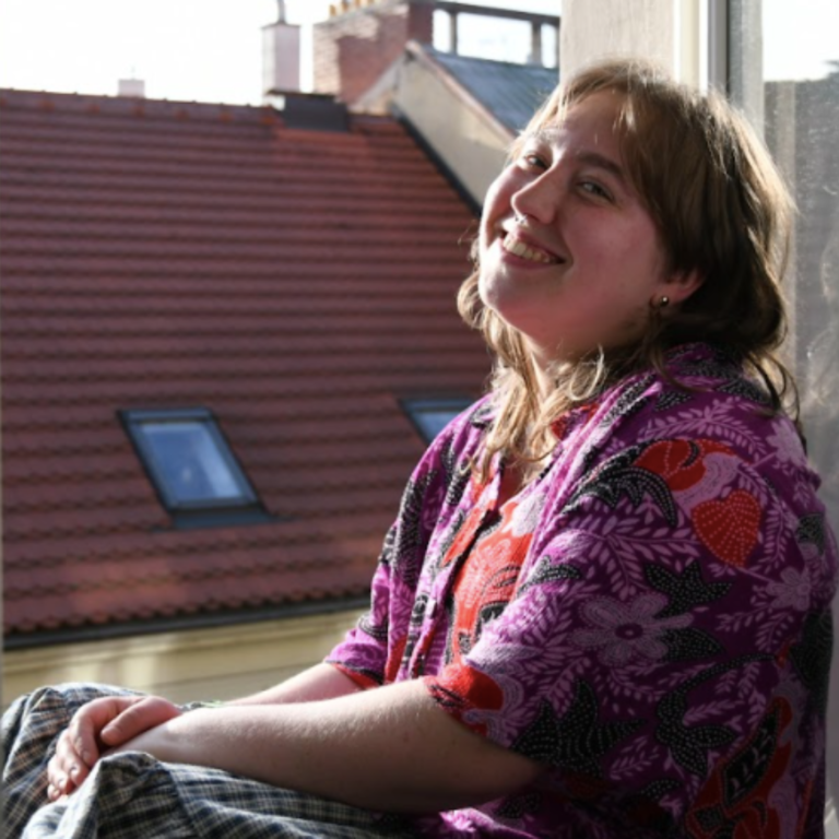 An image of a white nonbinary person with a purple blouse and wavy brown hair smiling at the camera. The background is the slanted roof of a Czech building.